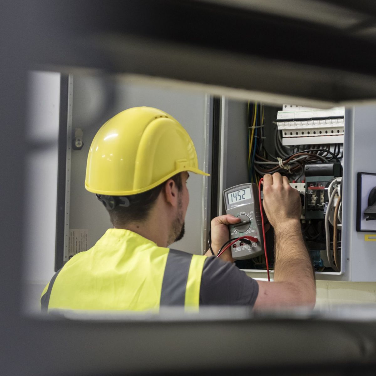 A technician wearing safety gear uses a digital multimeter to measure voltage within an industrial electrical control panel.