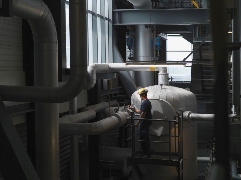 A worker stands on a platform, inspecting equipment in an industrial space filled with pipes and machinery.