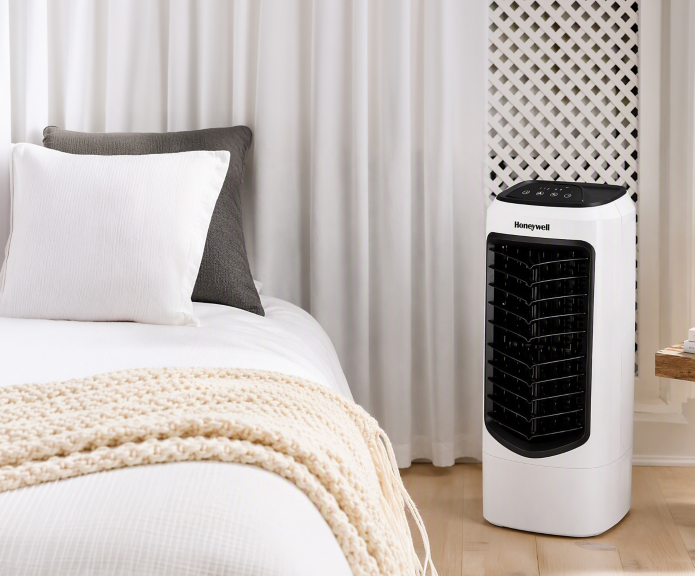 Honeywell evaporative air cooler with white housing and a black front grille positioned beside a bed in a bedroom.