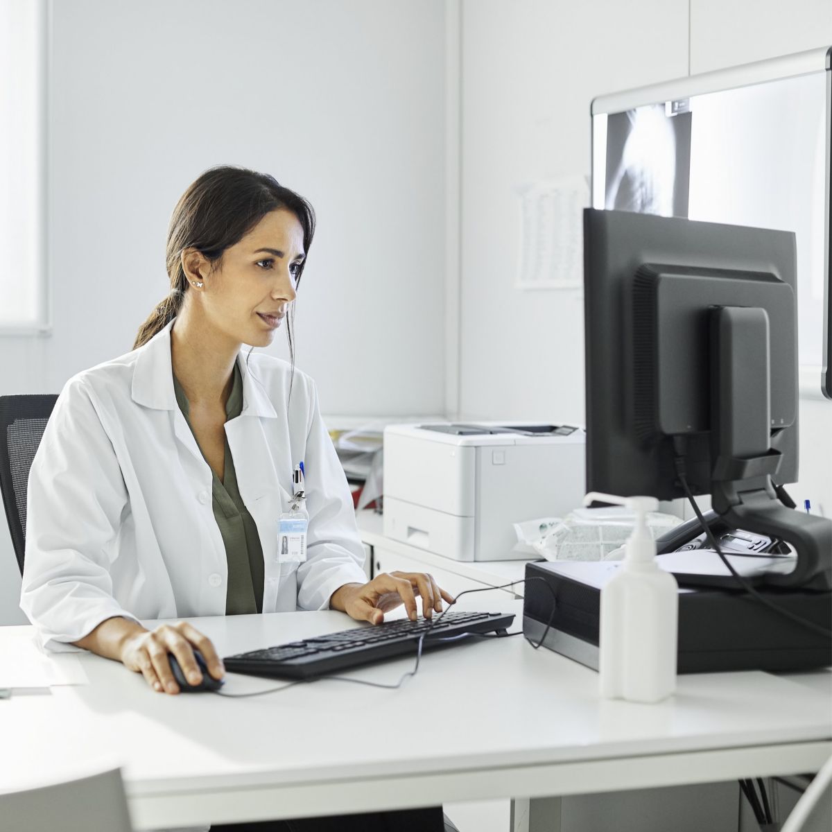 A professional female physician in a white coat works at her desk, using a computer in a clean, brightly lit hospital office environment.