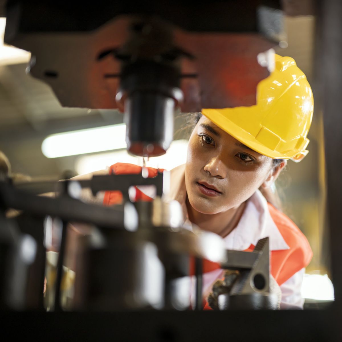A focused female engineer in a yellow hard hat carefully monitors industrial machinery during production.