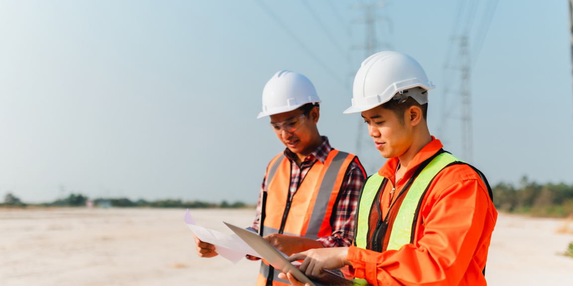 Two workers in safety gear reviewing documents at an outdoor site with industrial or utility infrastructure in the background.