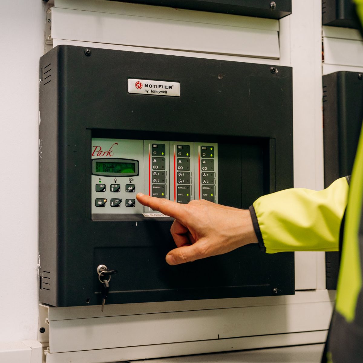 A technician in a high-visibility jacket operates a Notifier industrial fire alarm control panel.