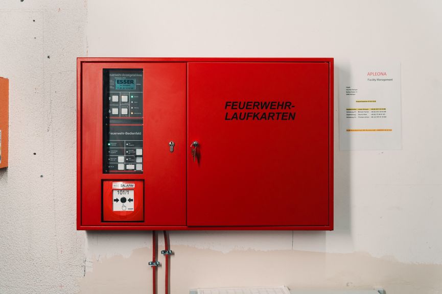 A red wall-mounted Esser fire alarm control panel with emergency map storage and contact information for facility management.