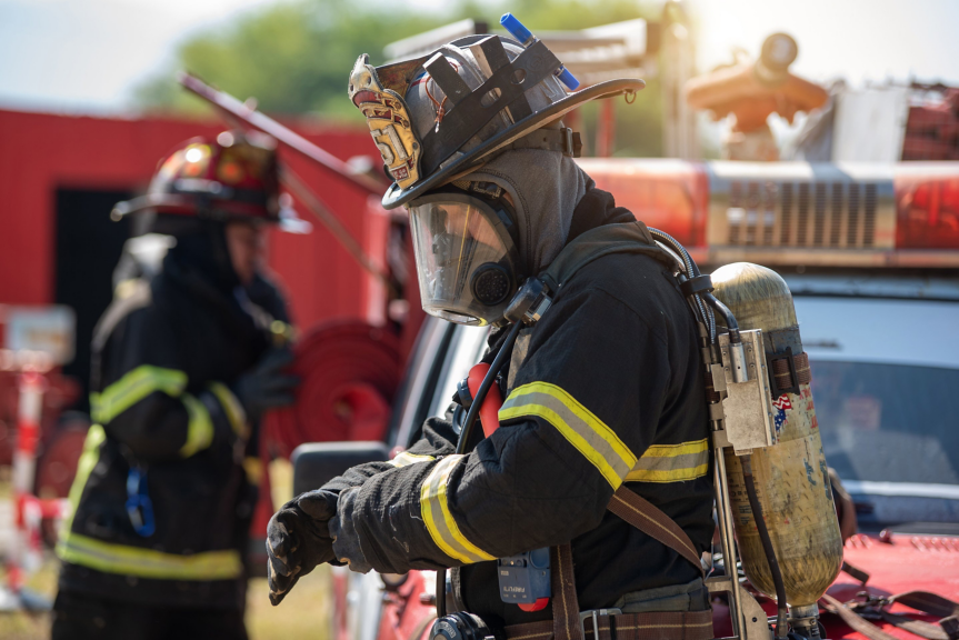 A firefighter in full protective gear adjusts their gloves in front of a fire engine during an emergency response training exercise.