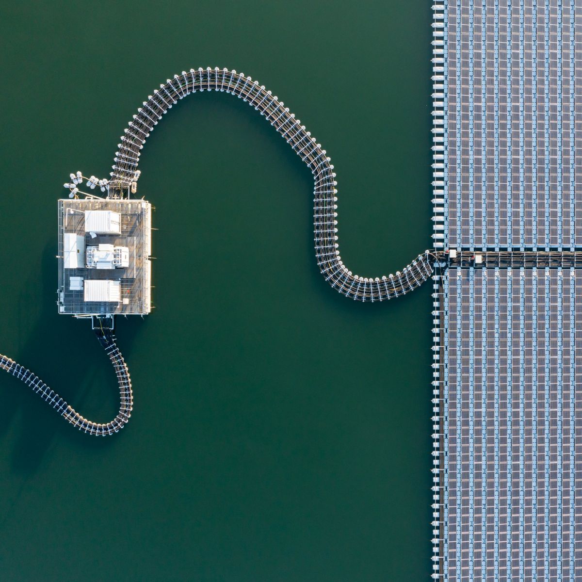 An aerial view displays a floating solar array connected to an electrical substation platform on a calm body of water.