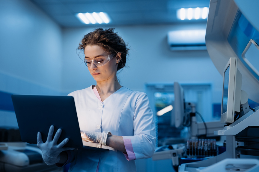 A healthcare worker in scrubs and gloves focuses on a laptop in a modern clinic, showcasing the vital role of technology in medical practice.