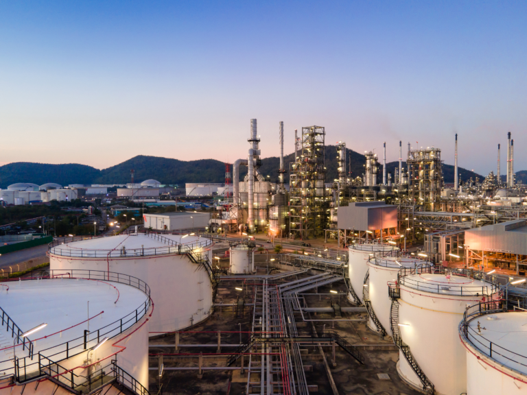Industrial refinery complex with cylindrical tanks, pipes, and illuminated towers under an evening sky.
