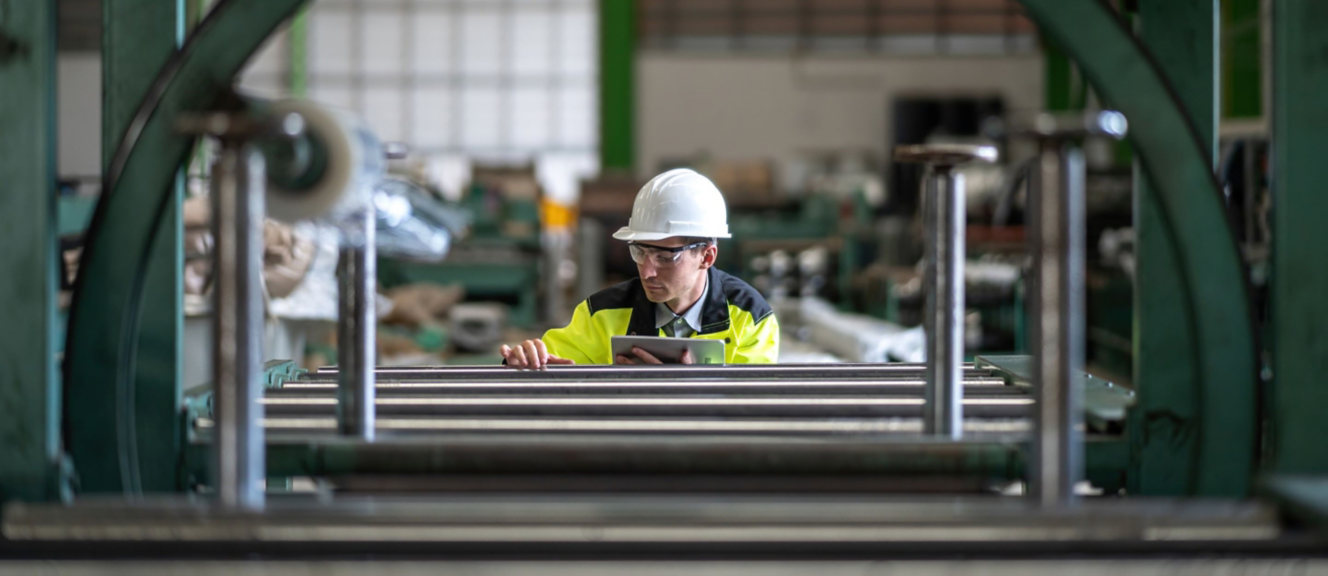 A factory worker in safety gear inspects machinery with a tablet, focused on operations in a busy industrial setting.
