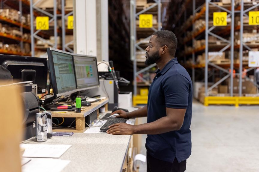 Side view of african male worker working on computer in a distribution warehouse. Man working on computer in large factory warehouse.