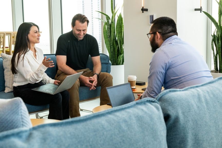 Three people sitting on chairs, one using a laptop, engaged in conversation in a room with plants, windows, and a small table.