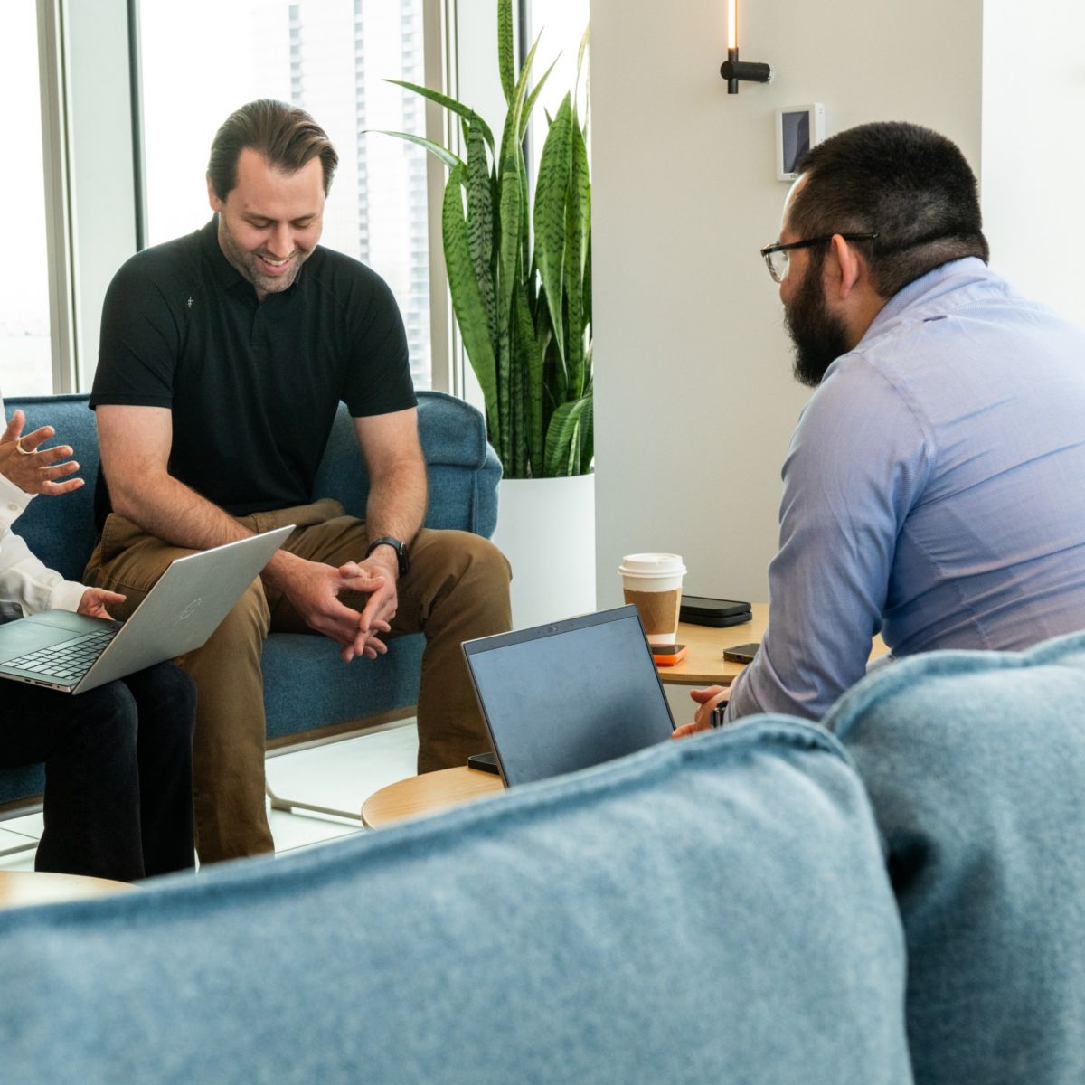 Three people sitting on chairs, one using a laptop, engaged in conversation in a room with plants, windows, and a small table.