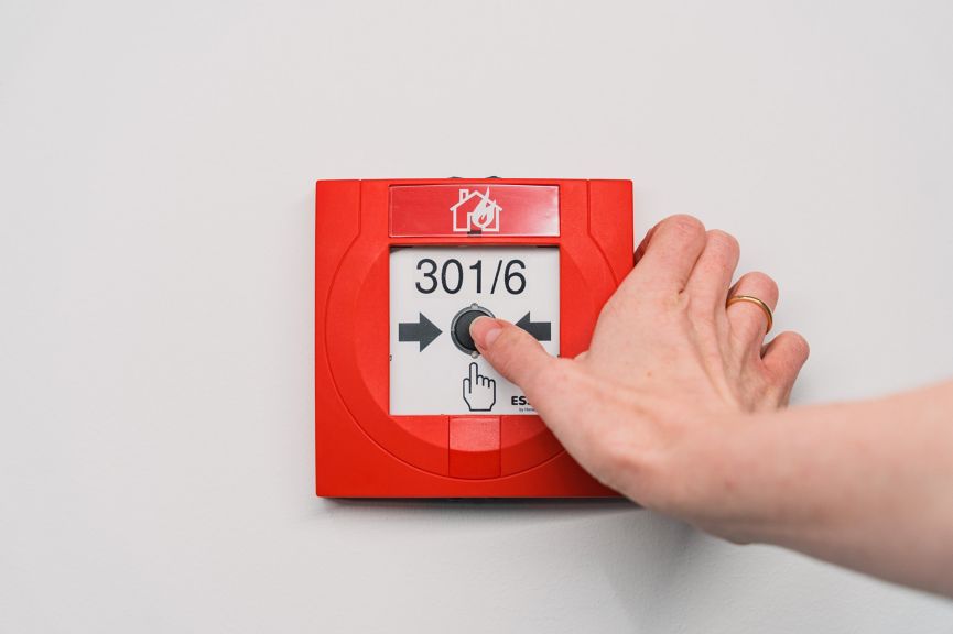 A person presses a red fire alarm call point button mounted on a white wall.