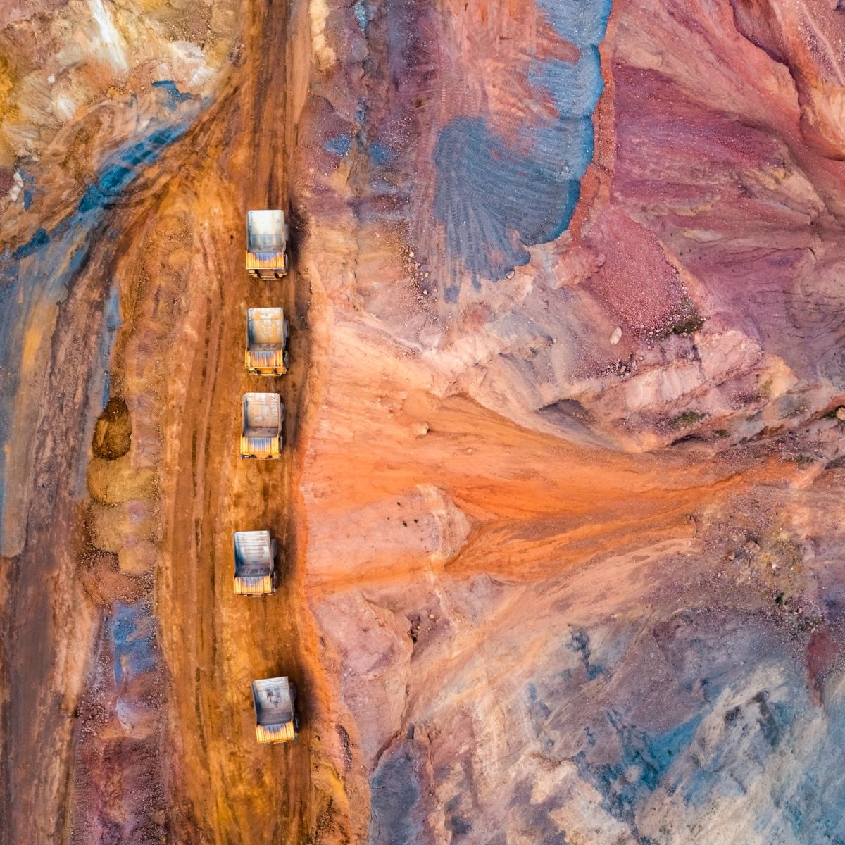 Aerial view of heavy-duty haul trucks driving in a line through a colorful, vibrant open-pit mining landscape.