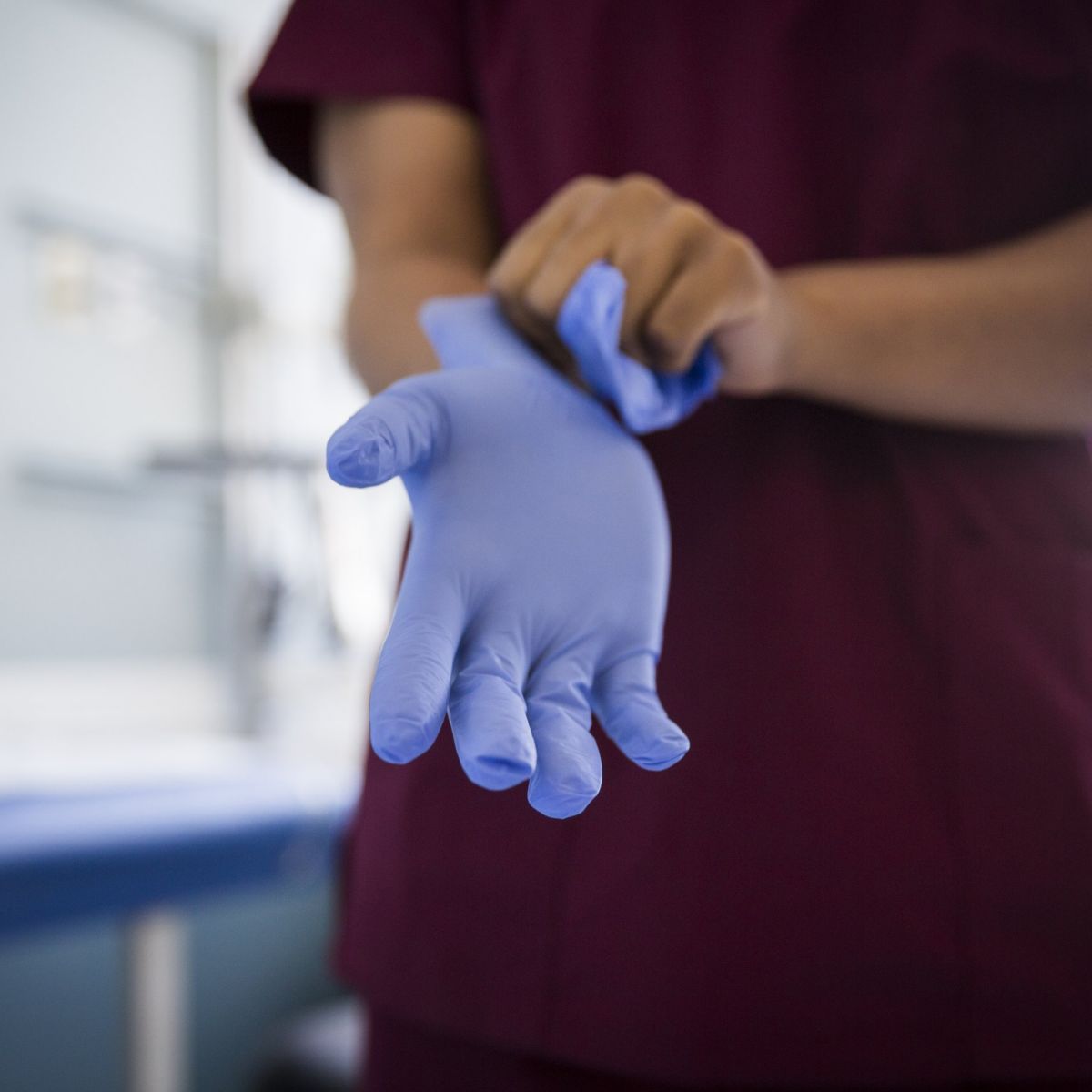 A healthcare worker in medical scrubs prepares for a procedure by carefully putting on protective blue nitrile gloves in a clinical setting.