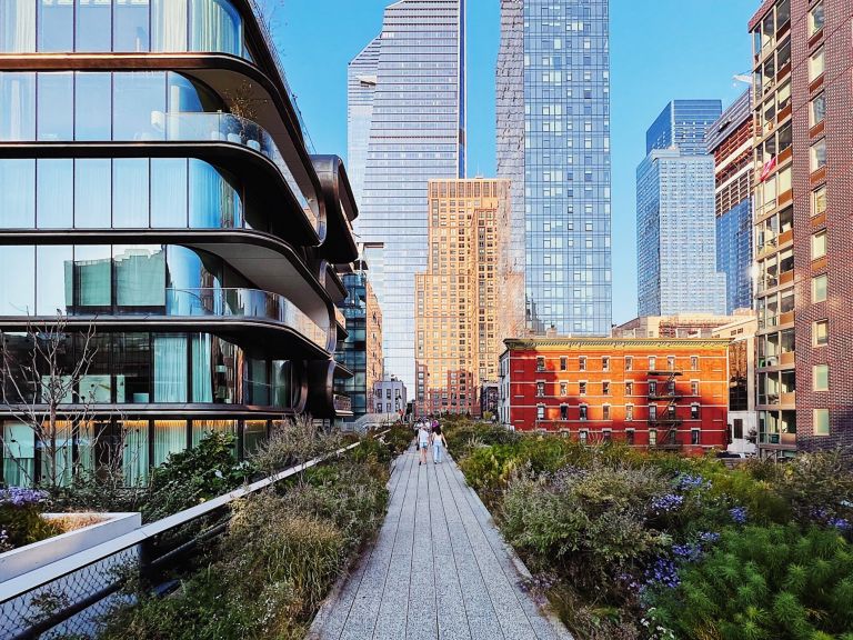 People walk along the High Line park in New York City, framed by Zaha Hadid's modern curves and traditional brick architecture under a clear blue sky.
