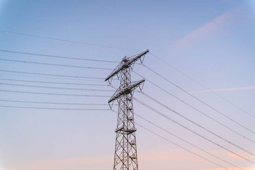 A tall metal transmission tower stands against a clear, soft-toned sky, showcasing complex electrical infrastructure in an industrial landscape.