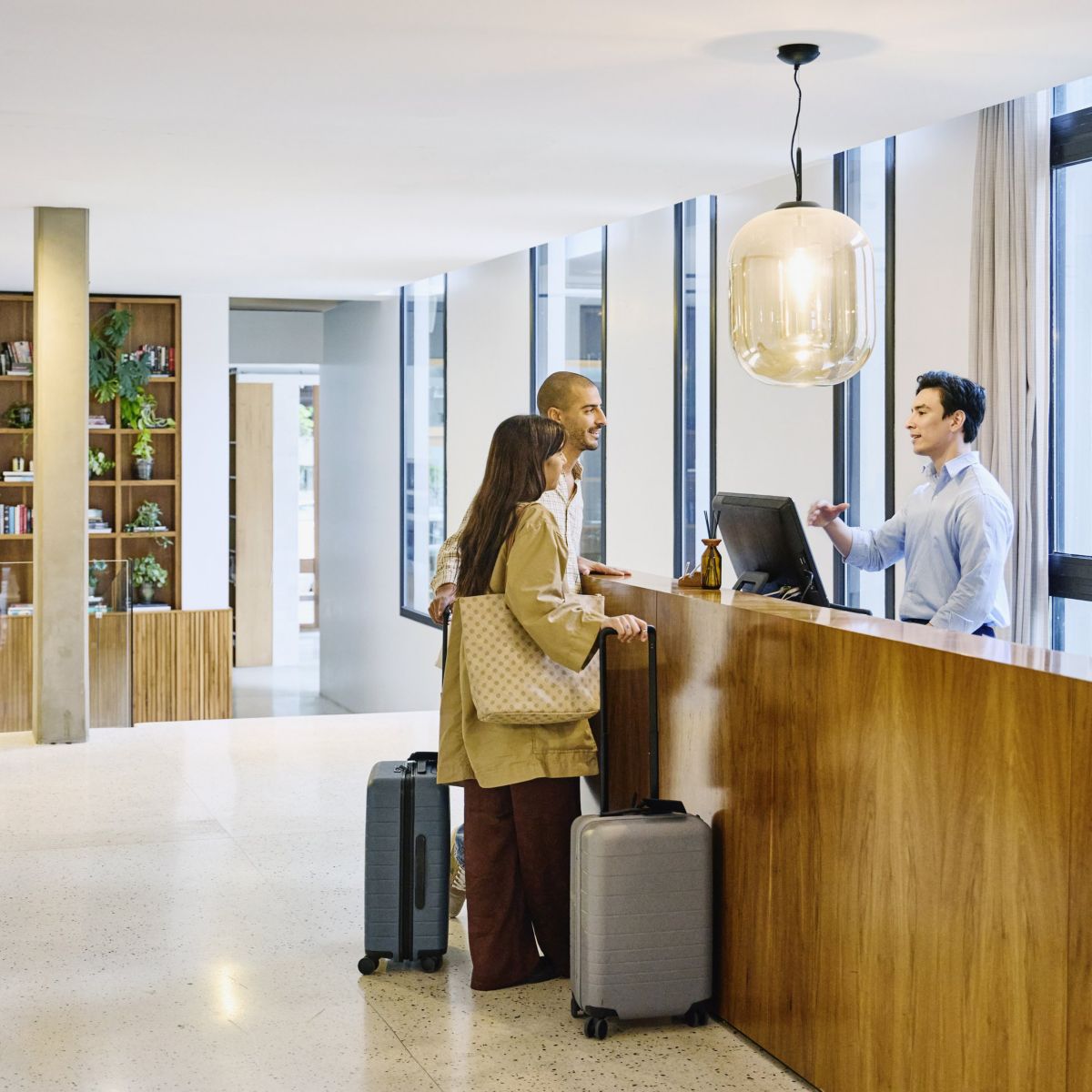 Guests engage with a hotel receptionist at a sleek wooden reception desk in a modern, well-lit lobby.