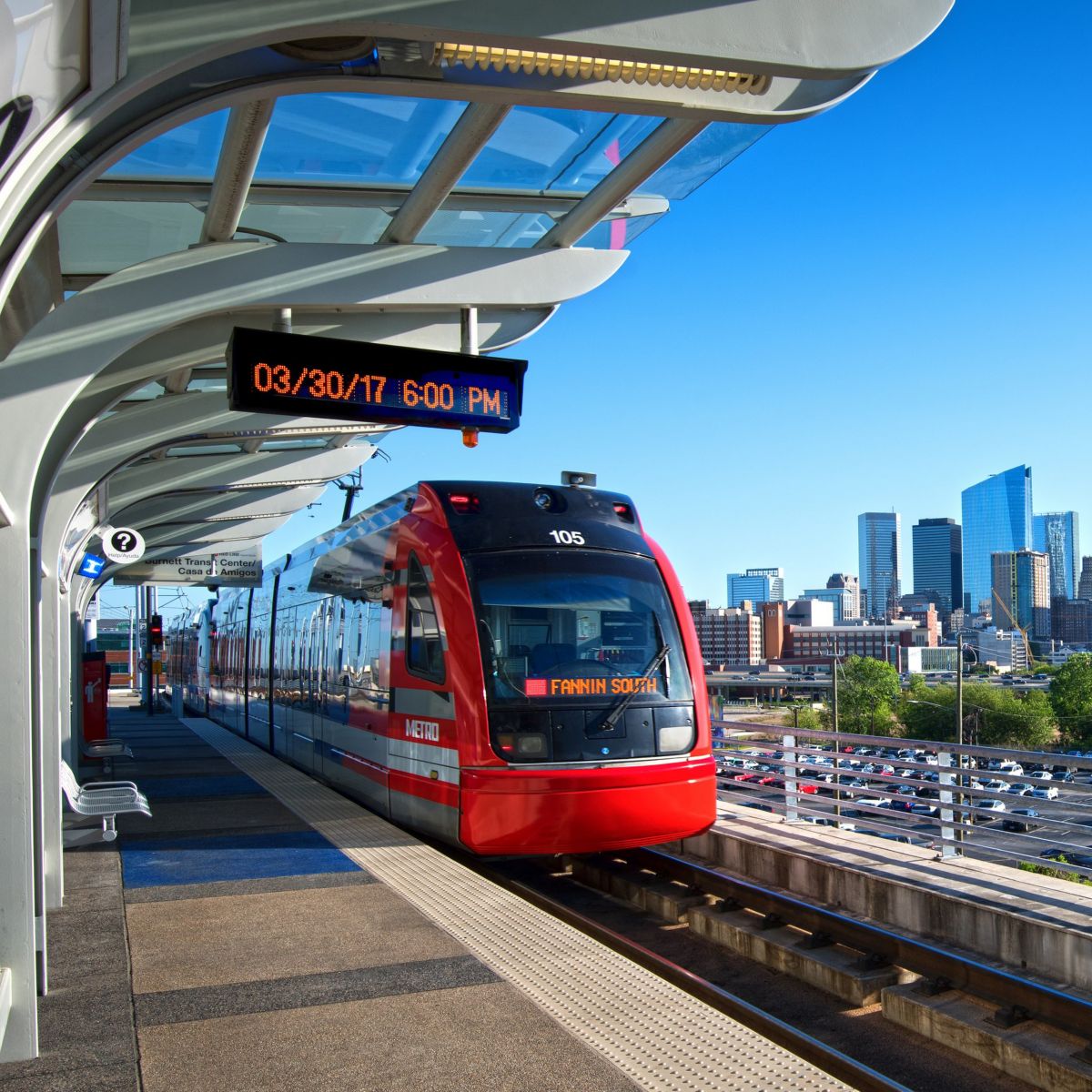 A METRO light rail train arrives at a modern elevated station with the Houston skyline visible in the background under a clear blue sky.