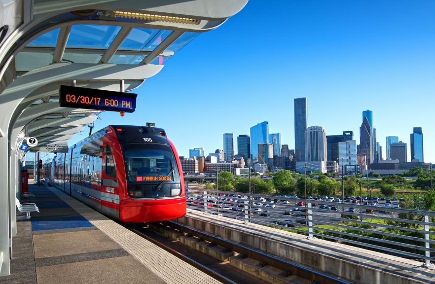 A METRO light rail train arrives at a modern elevated station with the Houston skyline visible in the background under a clear blue sky.