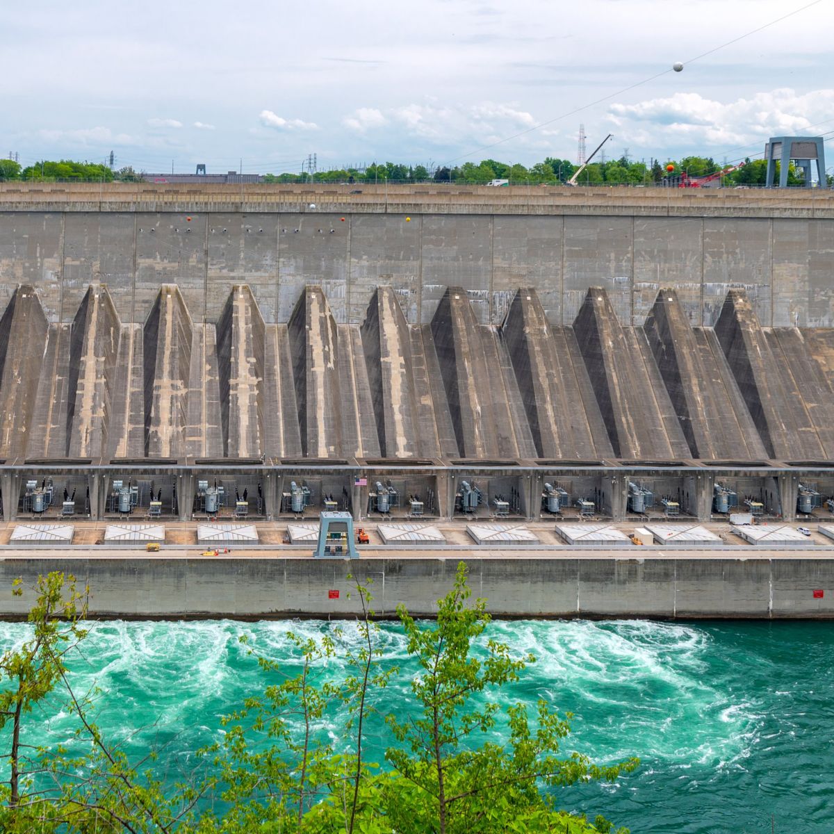 A wide view of a massive concrete hydroelectric dam structure showing its turbines, transformers, and the churning, turquoise water flowing below.
