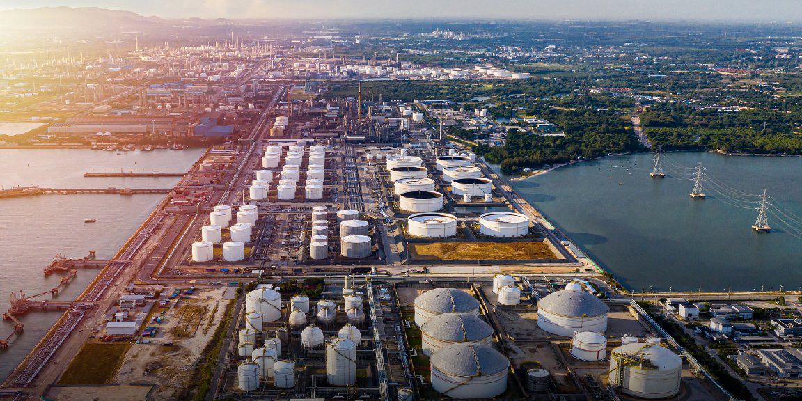 An industrial area with a large oil refinery featuring storage tanks, structures, terminals, and piping. Ships docked nearby suggest import/export activities. The intersection of industry, urban and natural landscapes is evident.