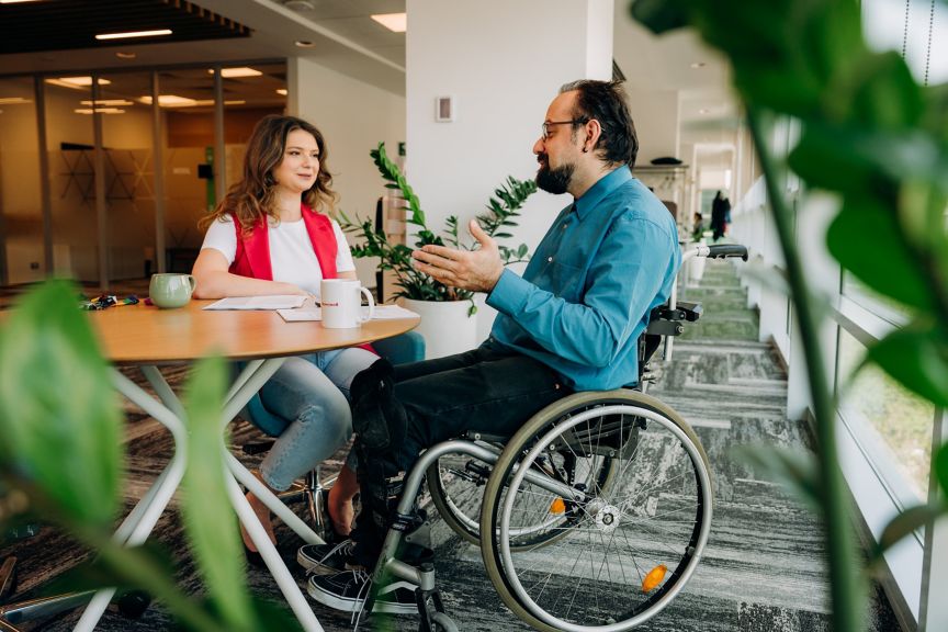 Two colleagues in a modern office engage in a professional discussion while seated at a round table, highlighting an inclusive corporate environment.