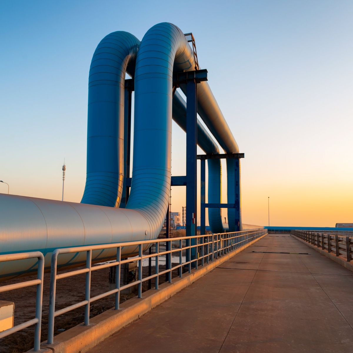 Large blue industrial pipes elevate over a concrete walkway against a clear sunset sky.