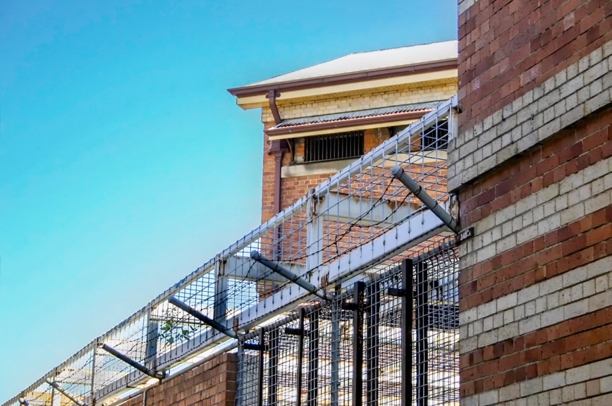 A perspective view of an industrial brick building secured by a tall metal mesh security fence under a clear blue sky.