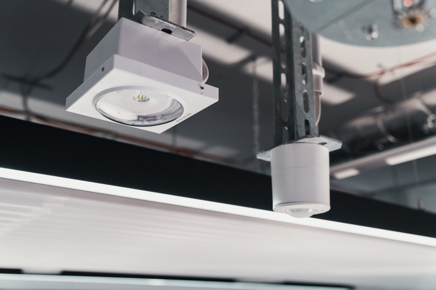 Industrial LED lighting and motion sensors are mounted to a suspended infrastructure system in an open-plan workspace.