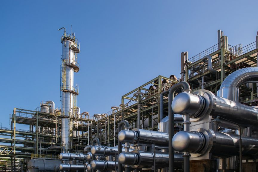 A complex array of insulated steel piping and distillation towers stands against a clear blue sky in an industrial chemical manufacturing facility.