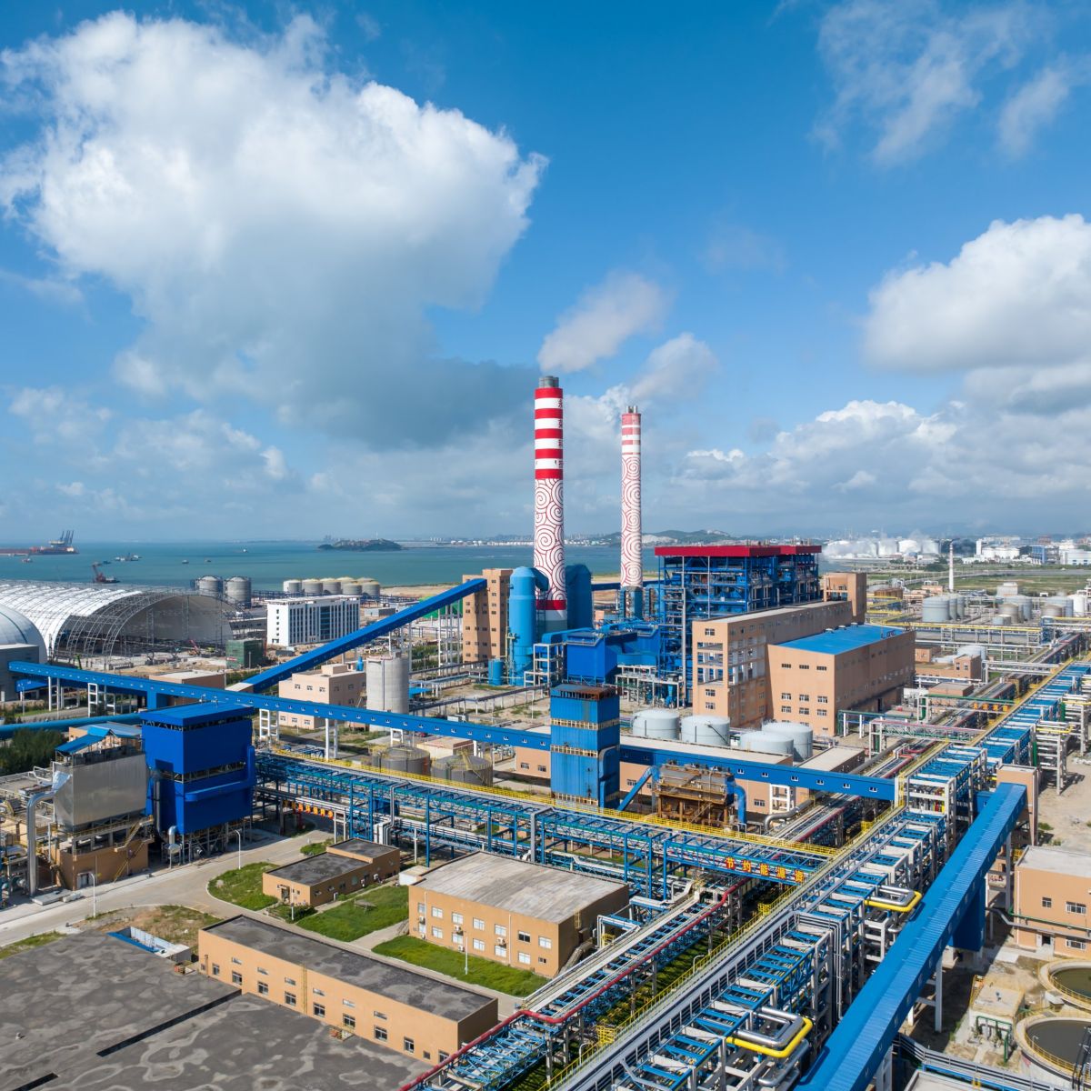 A high-angle aerial view of a sprawling coastal industrial facility featuring massive red-and-white striped chimneys under a vibrant blue sky.