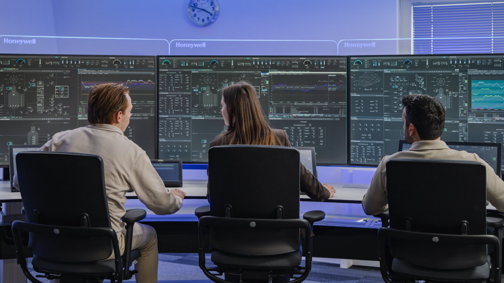 Three operators monitor complex data visualization screens in a high-tech corporate industrial control room environment.