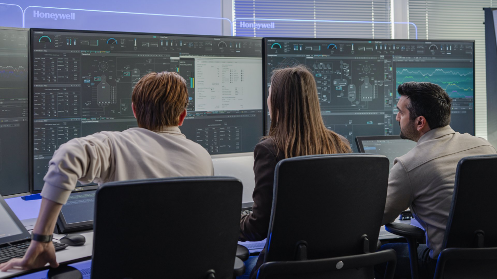 Three professionals analyze complex data and industrial workflows on large-scale digital displays in a modern Honeywell control room.