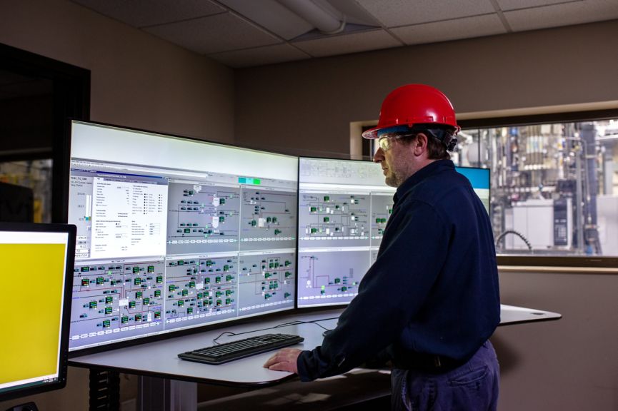 A worker in safety gear monitors complex industrial processes on multiple large screens within a control room setting.
