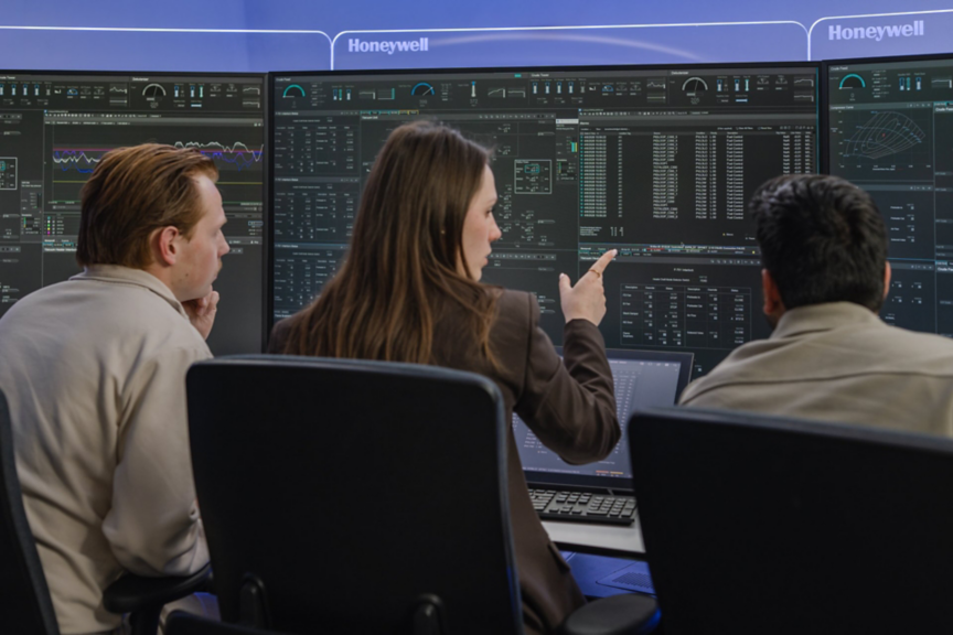 A team of engineers collaborates in a high-tech Honeywell control room, analyzing complex industrial data displays on a multi-screen workstation.