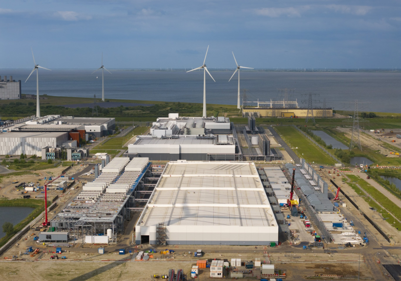 Aerial view of a large data center facility under construction, featuring multiple wind turbines and coastal energy infrastructure in the background.