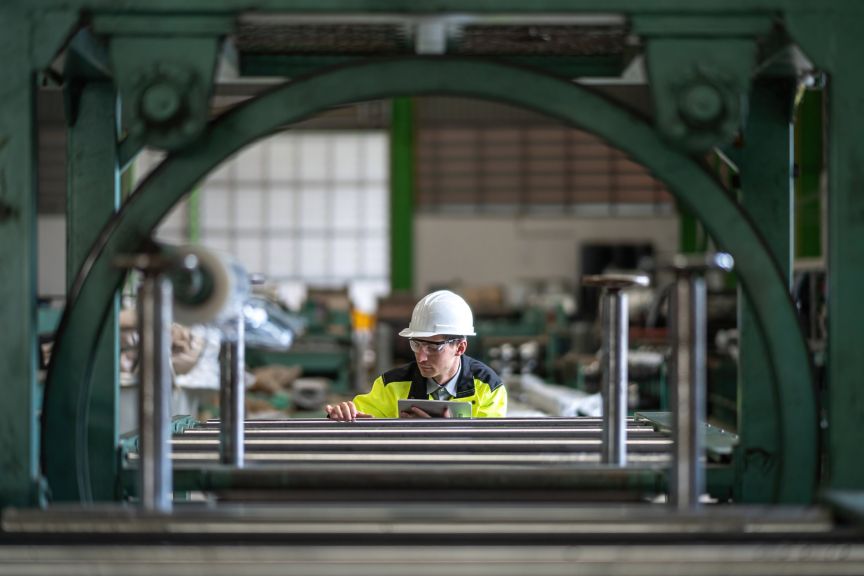 An engineer in protective gear reviews data on a tablet while inspecting machinery within a factory setting.