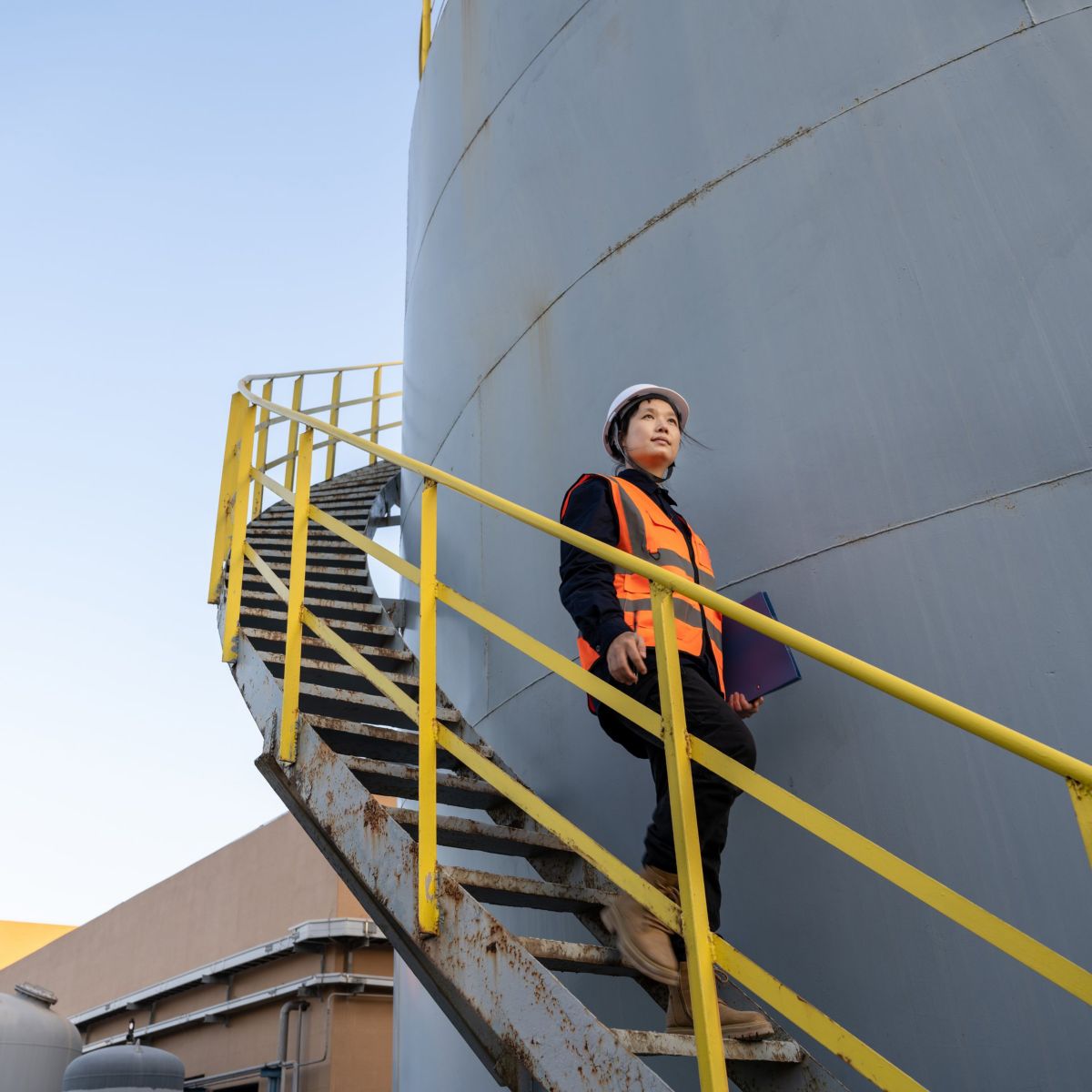 A female industrial engineer wearing a hard hat and safety vest walks down external stairs of a large storage tank while holding a blue clipboard.