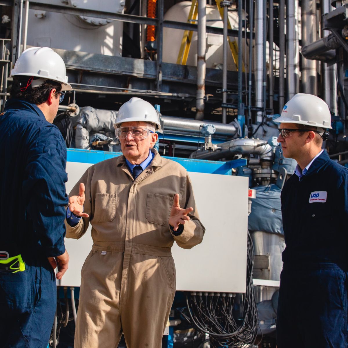 Three industrial professionals in hard hats and protective gear discuss operations in front of complex plant infrastructure.