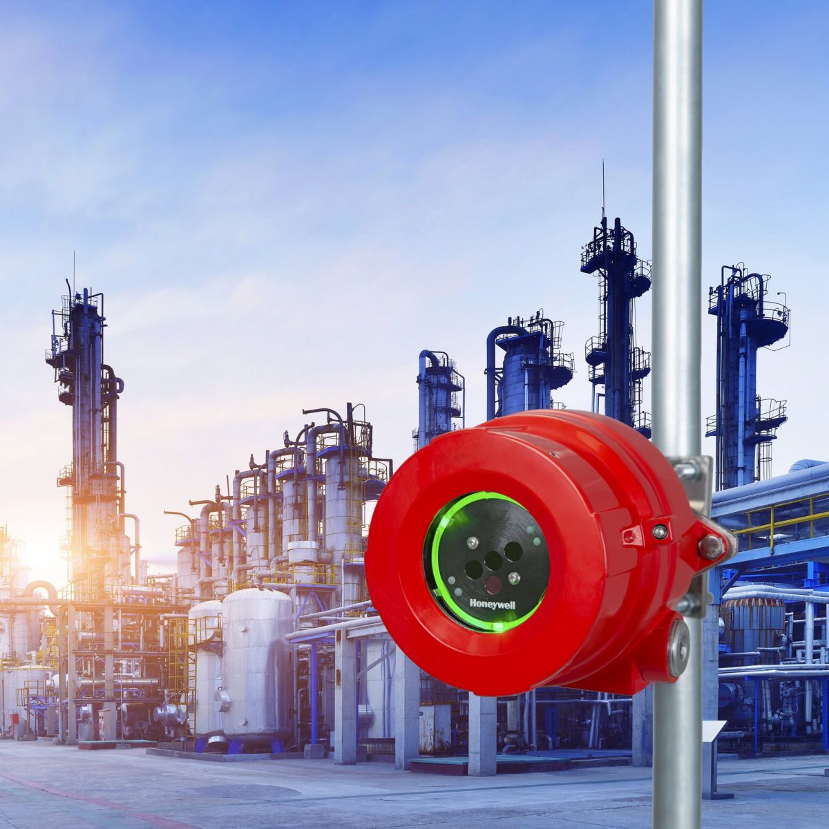 A bright red Honeywell gas detection sensor mounted on a pole against the backdrop of a petrochemical refinery facility at sunset.
