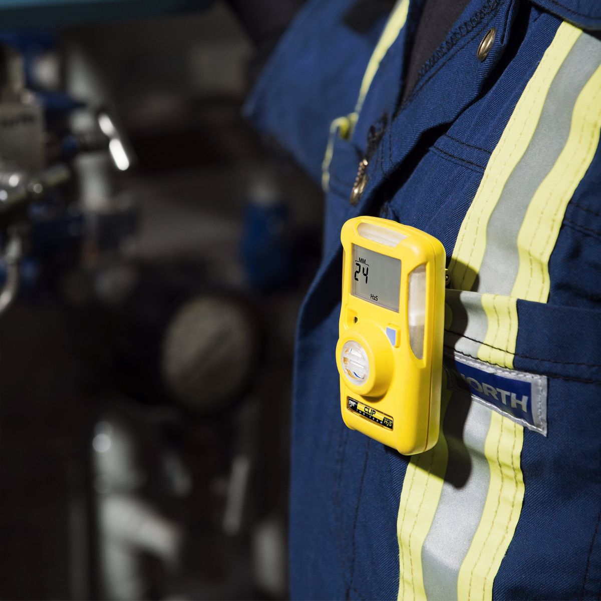 An industrial worker wears a personal hydrogen sulfide gas detector clipped to high-visibility work coveralls while operating equipment.