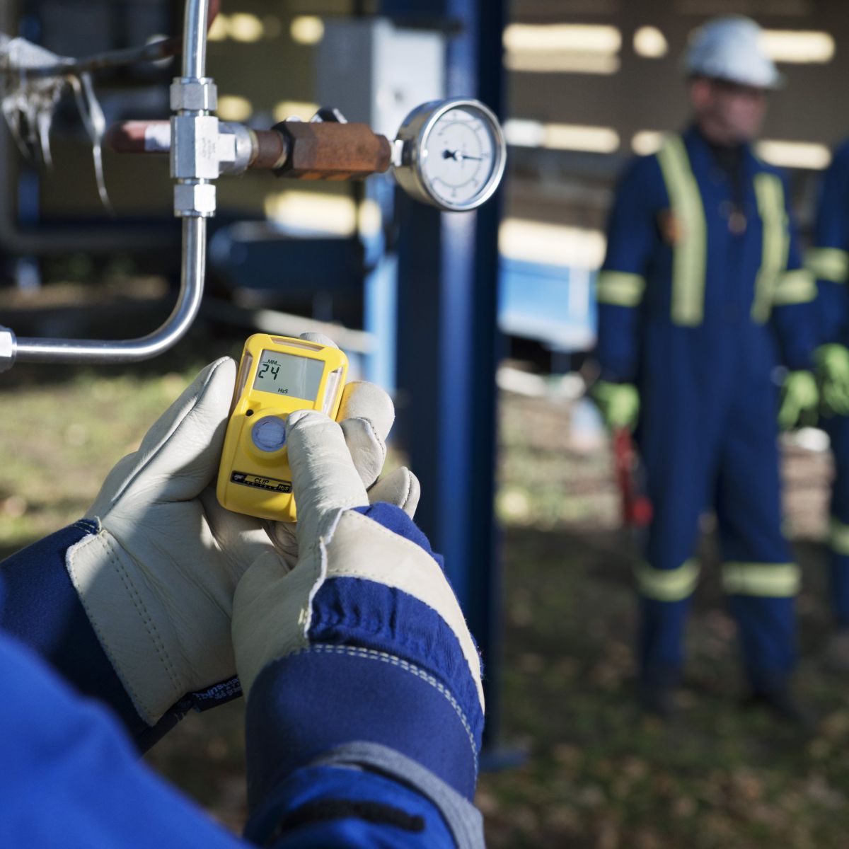 A worker in protective gear checks a portable gas detector near industrial piping, with colleagues standing blurred in the background.