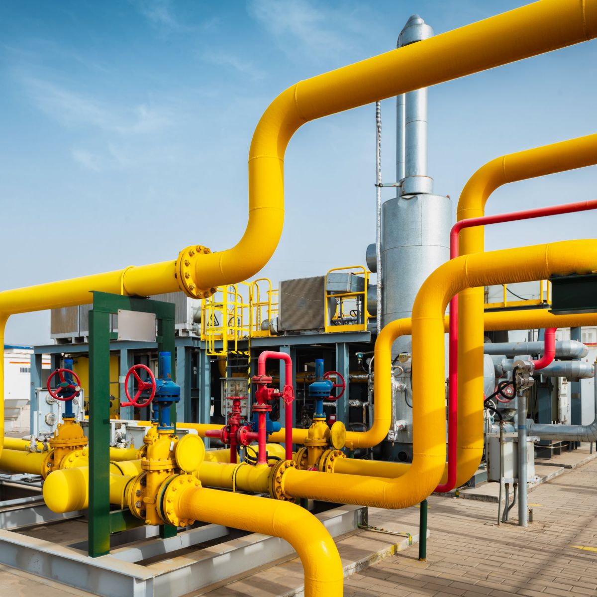 A vibrant network of yellow and red industrial pipes with valves at a natural gas processing facility under a clear blue sky.