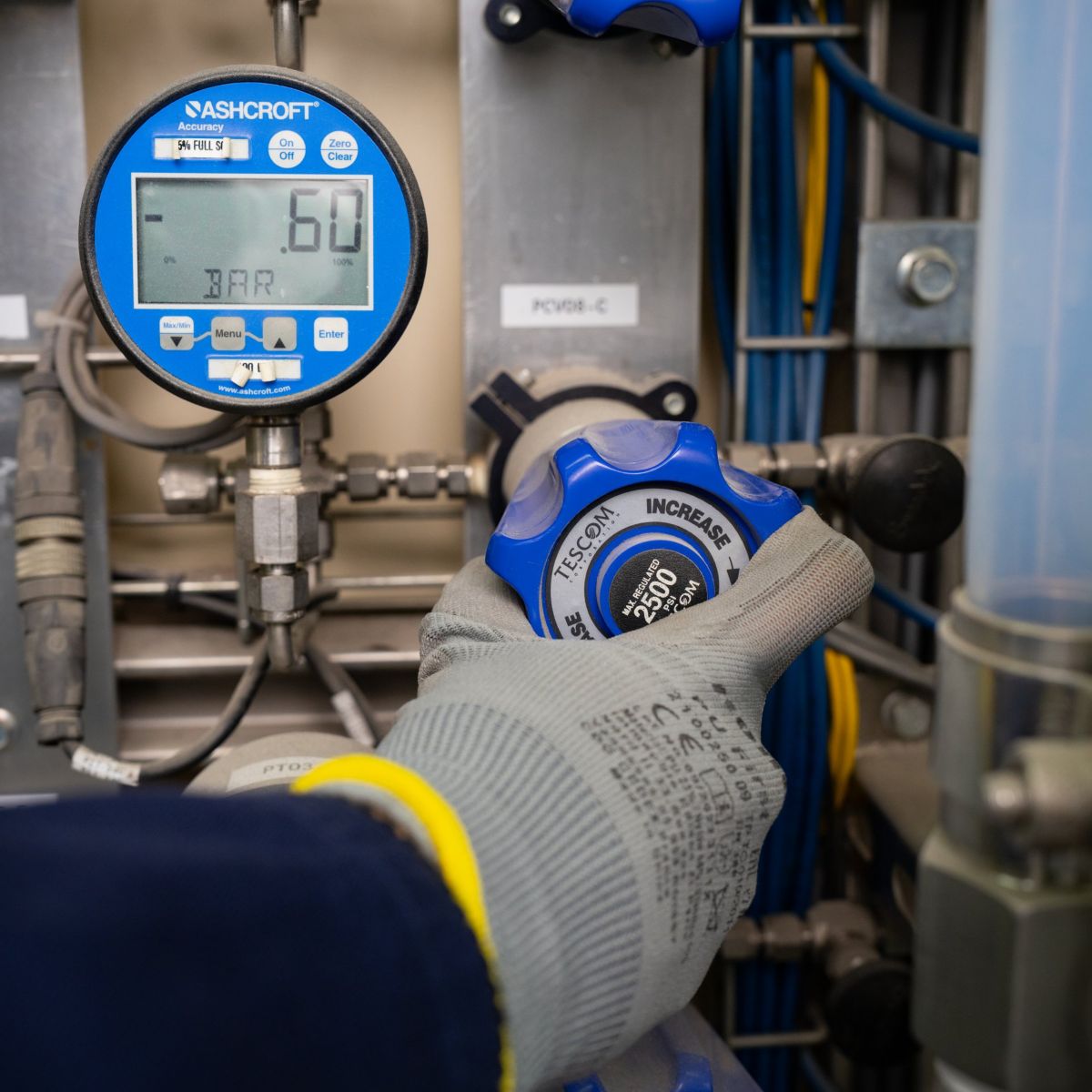 A gloved hand adjusts a Tescom pressure control valve next to an Ashcroft digital pressure gauge in a technical industrial environment.