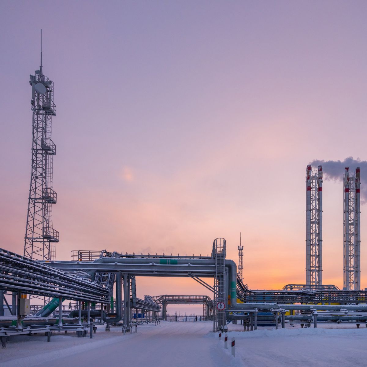 Pipes and steel towers at a natural gas plant are silhouetted against a vibrant, snow-covered sunset landscape during a cold winter evening.