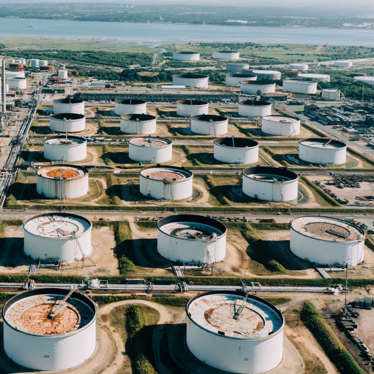 An aerial view shows an extensive oil refinery complex featuring large storage tanks and processing infrastructure near a coastal landscape.