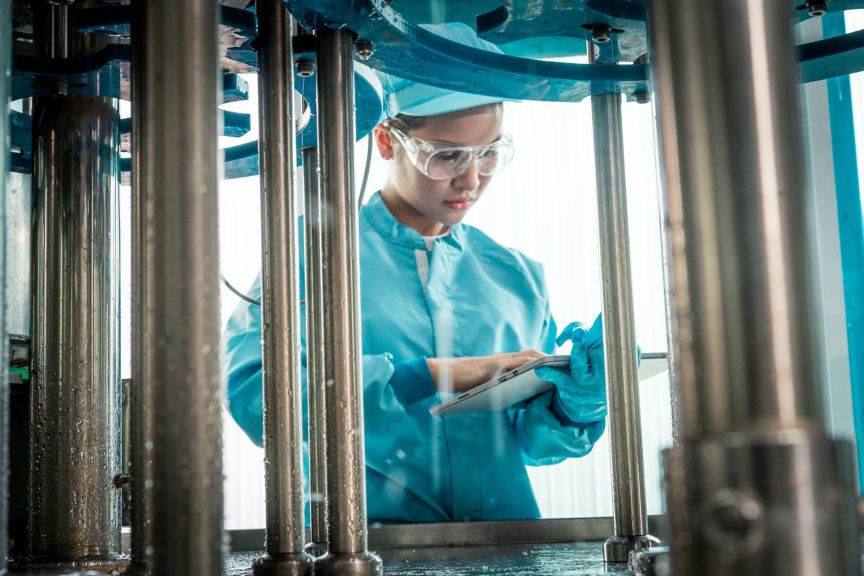An operator in protective gear monitors industrial machinery using a tablet inside a beverage bottling facility.
