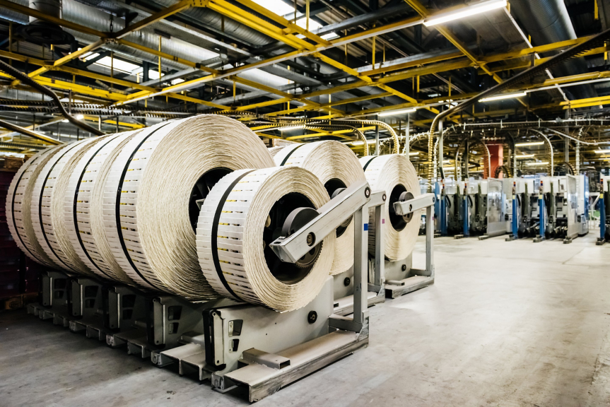 Large industrial paper rolls are staged on automated stands within a modern manufacturing facility featuring overhead yellow steel structural supports.