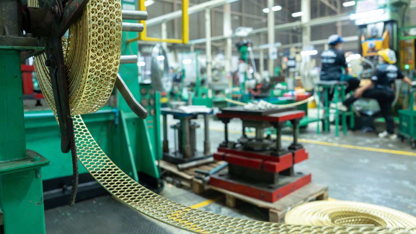 A close-up view of a perforated brass coil feeding into heavy manufacturing machinery within a bustling factory setting.